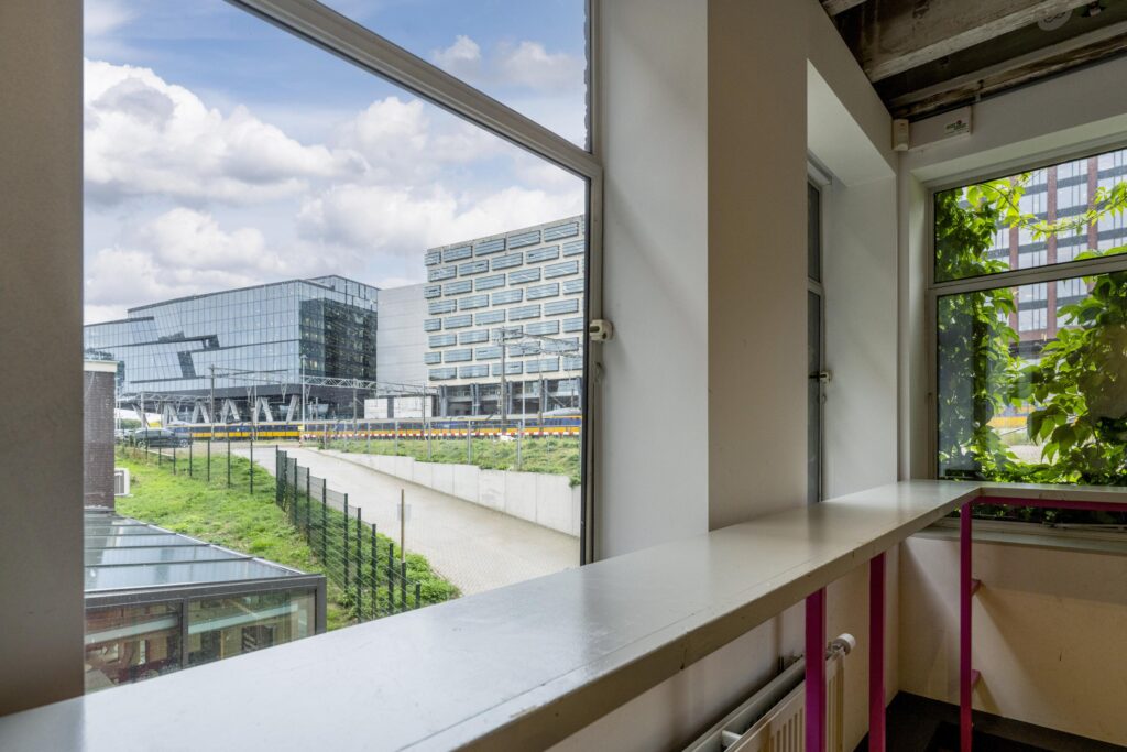 Uitzicht vanuit een interieur aan de De Ruijterkade op moderne kantoorgebouwen en het treinspoor bij station Amsterdam Centraal.